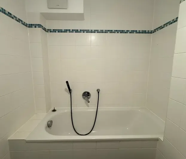White bathtub with a handheld shower on a white-tiled wall, accented by a blue mosaic border in a modern bathroom.