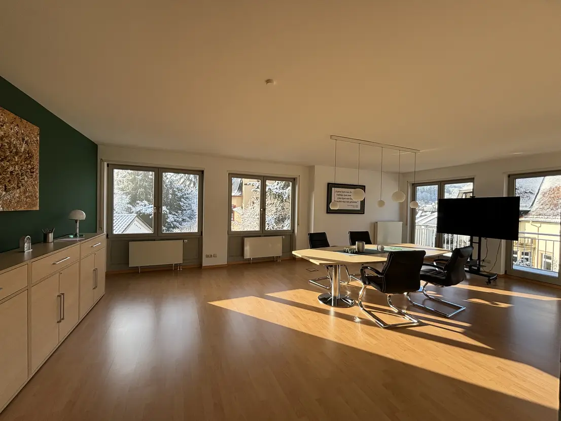 Bright, modern conference room with a long white table, black swivel chairs, and a wall-mounted TV by large windows overlooking rooftops misted by sunlight.