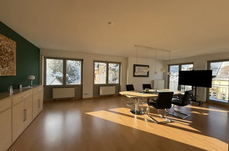 Bright conference room with a long table, black swivel chairs, and a large wall-mounted TV, sunlit by windows to the outside.