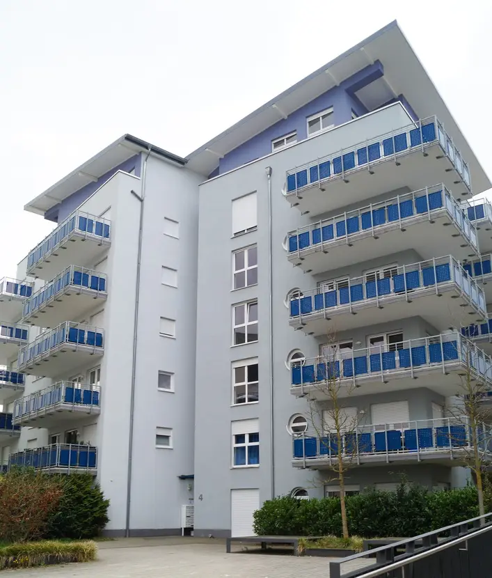 Modern gray apartment building with multiple blue-railed balconies and a paved courtyard area.