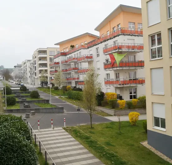 Residential apartment complex with white buildings and red balconies, flag on the front building, beside a pedestrian path and green lawn.