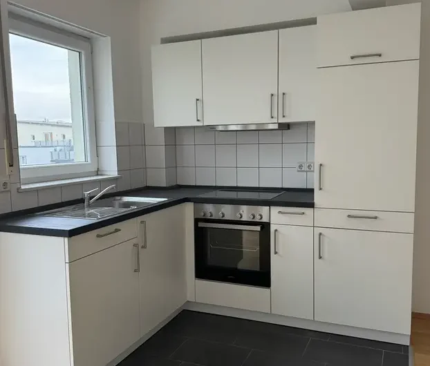 Compact white L-shaped kitchen with built-in oven, electric stove, stainless sink, and white cabinets; window to the left and dark countertops.