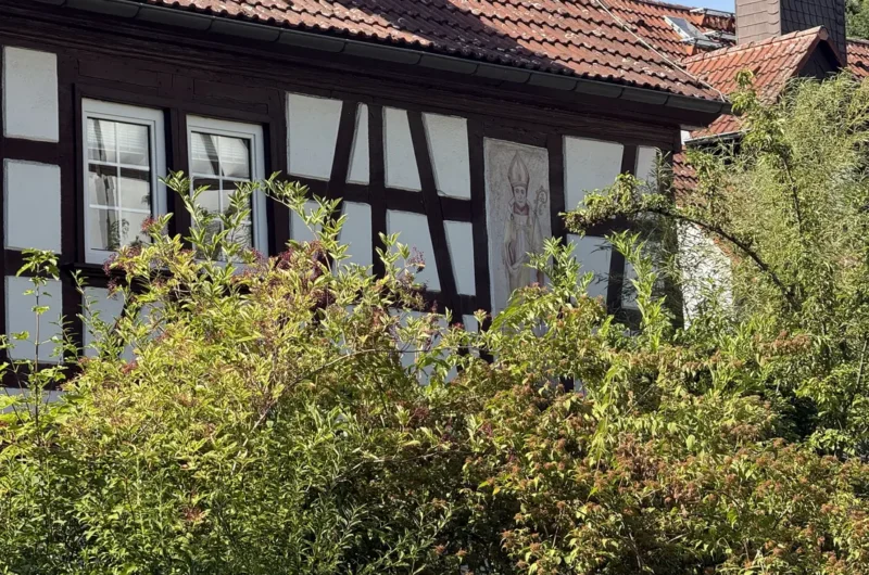 Timber-framed house with white walls and dark beams, two windows, red-tiled roof, and garden shrubs in front.