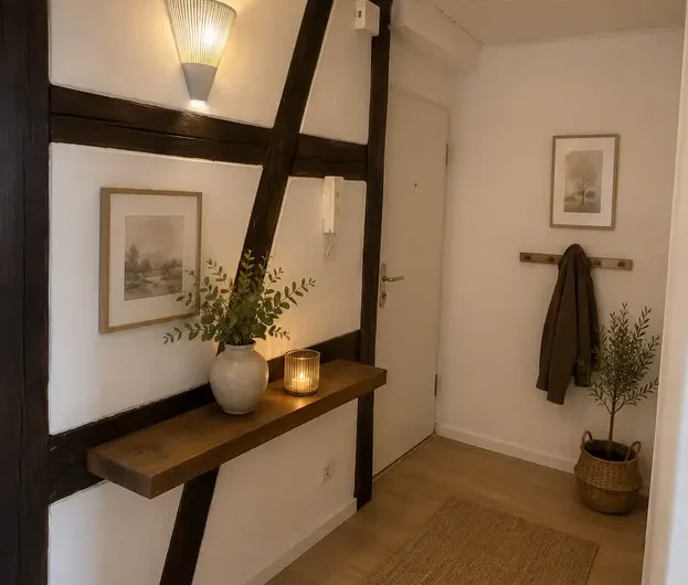Entry hallway with dark wooden beams, a narrow wooden console shelf holding a vase of greenery and a lit candle, framed artwork, and a coat on a wall rack near the door.