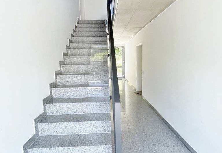 Indoor stairwell with gray terrazzo steps and a black metal handrail, running alongside a white-walled hallway to a doorway at the far end.