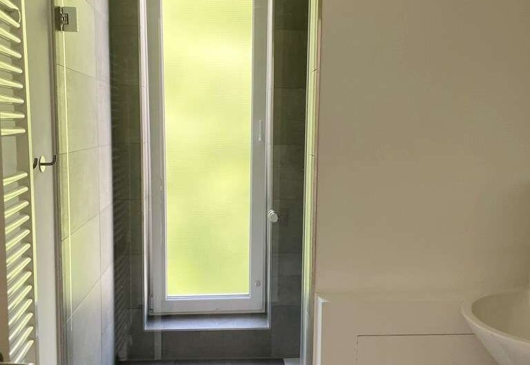 Modern bathroom with a glass-walled shower stall, tall frosted window, and gray tile walls; white vanity on the right and a vertical towel radiator on the left.