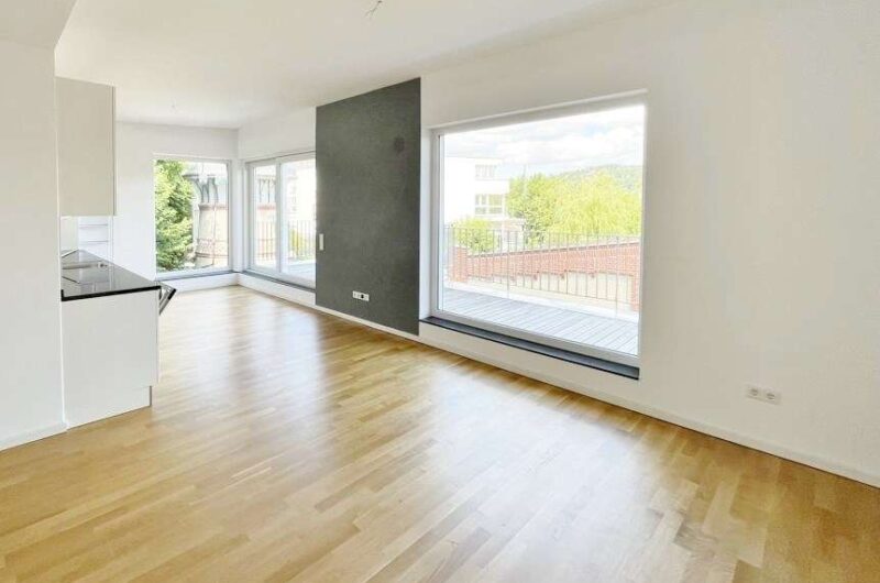 Bright open living space with hardwood floors, large windows, and a dark accent wall; a small white kitchen on the left and a balcony outside.