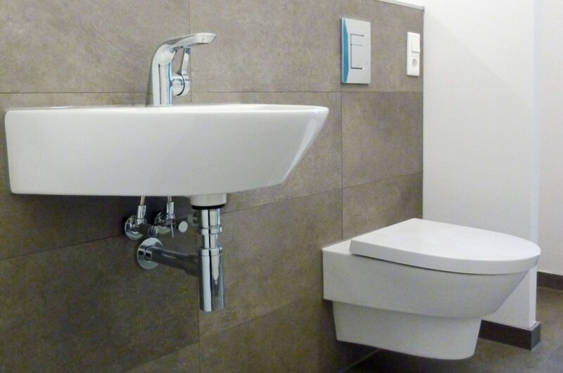 Modern bathroom with a wall-mounted white rectangular sink and exposed chrome pipes beside a wall-mounted toilet on grey tiled walls.