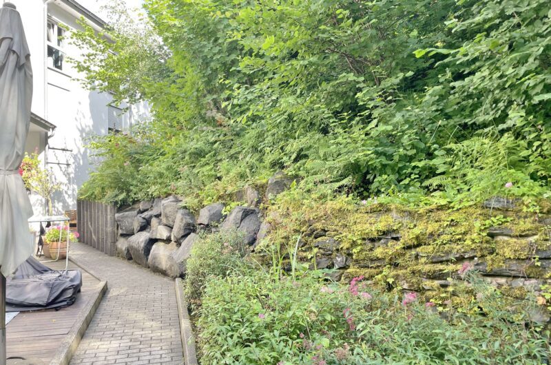 Narrow stone terrace beside a lush green hedge and a white house on the left, under bright daylight with a covered outdoor area nearby.