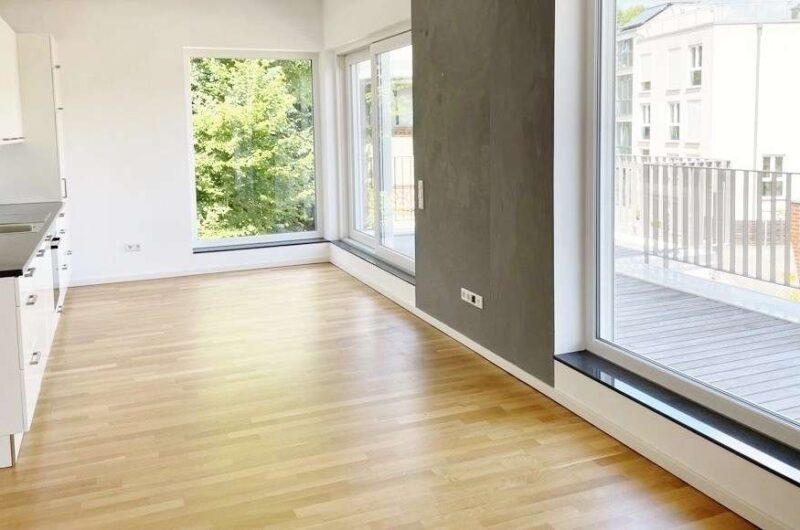 Bright empty living area with wide floor-to-ceiling windows, light hardwood floors, and a white kitchen on the left.