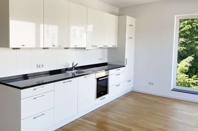 White modern kitchen with glossy cabinets, black countertop, sink, and built-in oven under a windowed wall with greenery outside