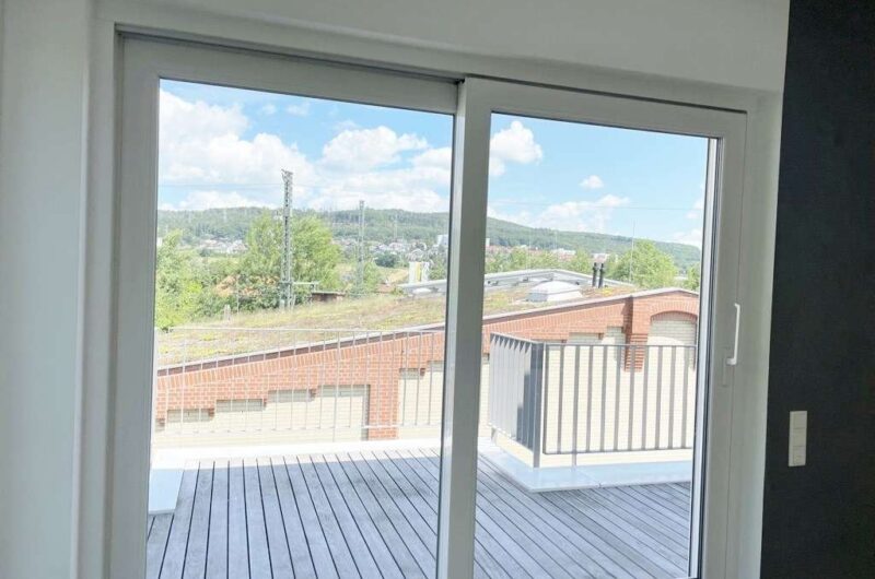 Sliding glass doors open to a wooden deck with a railing, overlooking brick rooftops, trees, and a green hillside under a blue, cloudy sky.