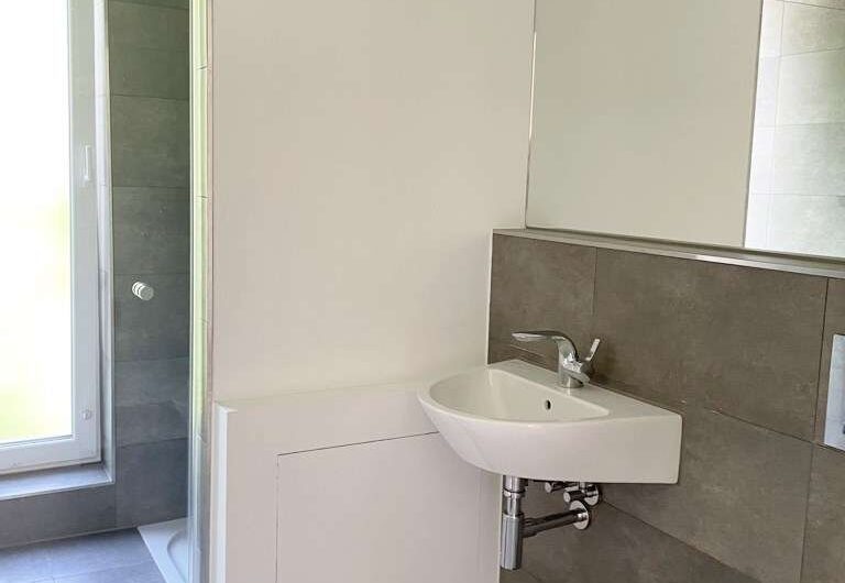 Modern bathroom with a wall-mounted white sink, chrome faucet and exposed plumbing, grey tile walls and floor, a large mirrored cabinet, and a glass-door shower to the left by a window. Showers and toilet partially visible in a compact layout.